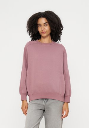 Kvinde med krøllet hår iført en løs støvet lyserød sweatshirt og lysegrå jeans, stående foran en enkel lys baggrund.