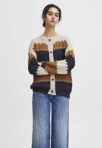 Austs adīts kardigans ar zigzaga rakstu krēmā, brūnā un jūras zilā krāsā. Tam ir apaļa kakla izgriezums, pogu aizdare, kā arī ribota aproce un apakša.