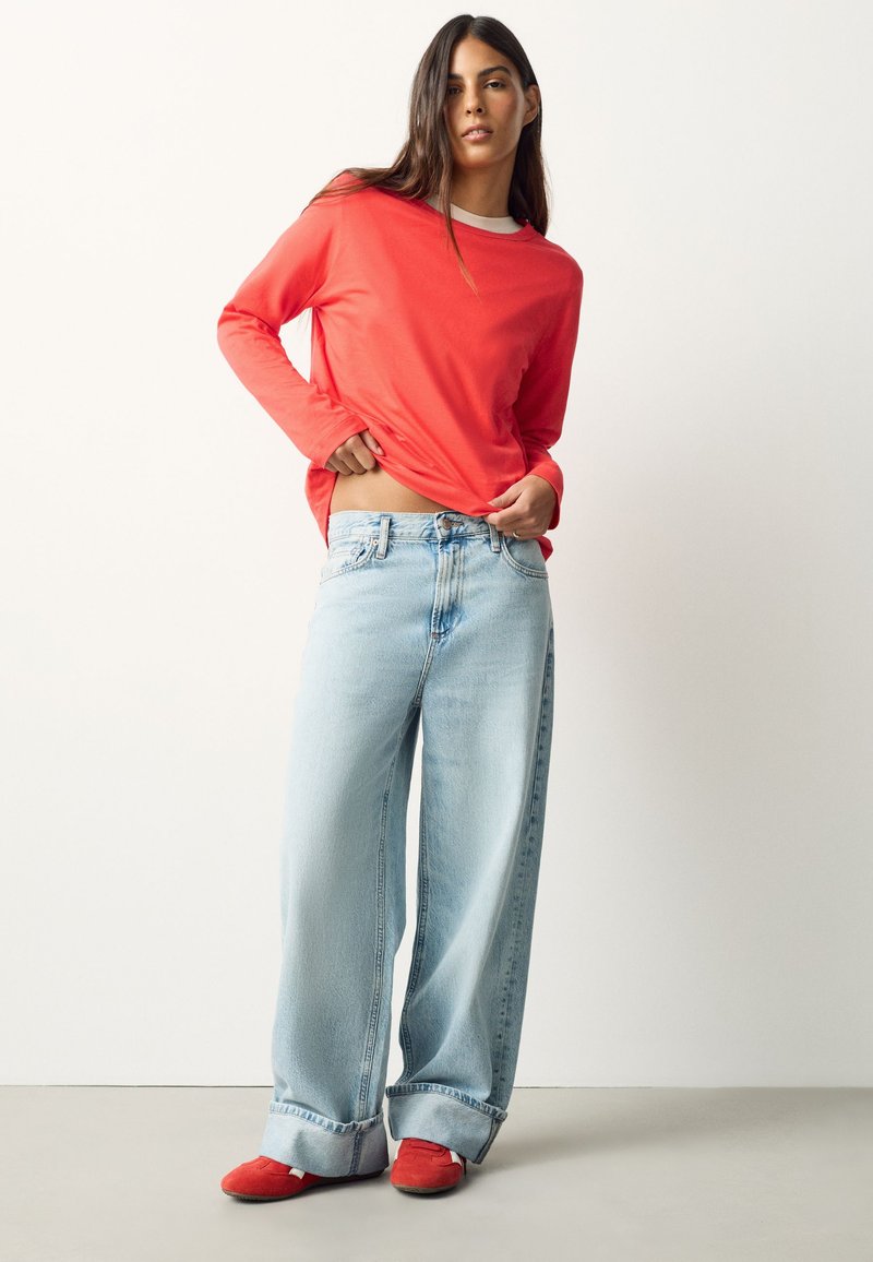 Donna con camicia rossa a maniche lunghe, jeans larghi azzurri chiari e scarpe rosse, in piedi davanti a uno sfondo bianco uniforme.