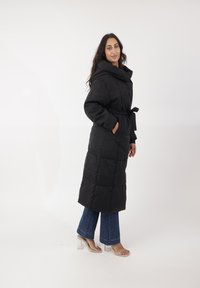 Cappotto imbottito nero con un ampio cappuccio, vita con cintura, due tasche laterali e una silhouette lunga e dritta. Indossato sopra jeans a zampa d'elefante blu e tacchi trasparenti.