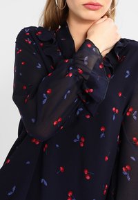 Donna che indossa una blusa blu navy con un motivo di ciliegie rosse e colletto arricciato, mano che tocca il collo, grande orecchino a cerchio visibile.