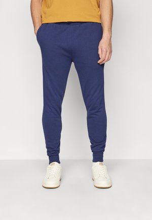 Mann trägt dunkelblaue Jogger-Hosen mit Taschen, kombiniert mit weißen Sneakers und einem senfgelben Hemd, steht vor einem einfachen Hintergrund.