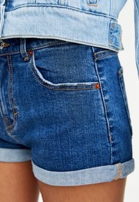 Primer plano de una persona que lleva pantalones cortos de mezclilla oscuros arremangados y una chaqueta de mezclilla de color azul claro.