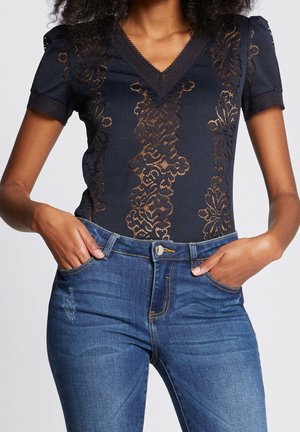 Mujer con top azul de encaje ajustado con patrones florales y vaqueros azules, con las manos metidas en los bolsillos delanteros.