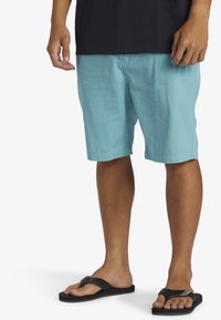 Ljust blå bomullsshorts med en avslappnad passform och sidofickor, parat med svarta flip-flop-sandaler på barfot. Slät textur och enkel design.