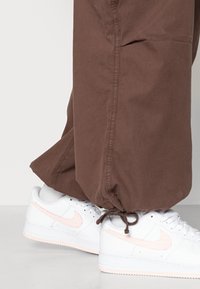 Bílé tenisky Nike s růžovými akcenty, v kombinaci s volnými hnědými kalhotami na šňůrku. Kalhoty mají strukturovaný povrch a elastické manžety.