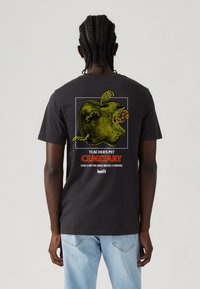 Person med fletninger iført en sort t-shirt med et grønt råddent æble med orme og teksten "Teacher's Pet Cemetary" og "Levi's."