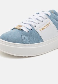 Baskets en suède bleu clair avec des accents en cuir blanc, présentant un logo doré sur le côté et des lacets blancs, montées sur une semelle en caoutchouc blanche.