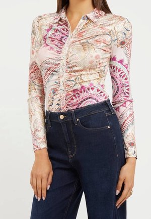 Kvinde iført en langærmet skjorte med lyserøde og beige paisley-mønstre, kombineret med højtaljede mørkeblå jeans.