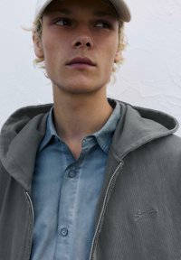 Grauer Reißverschluss-Hoodie mit strukturierter Oberfläche, getragen über einem hellblauen Button-Down-Hemd mit zwei Knöpfen, vor einem weißen Hintergrund präsentiert.