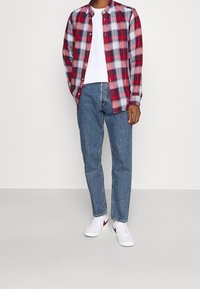 Rode en blauwe geruite button-up shirt over een wit T-shirt, gecombineerd met lichtblauwe jeans en witte sneakers met een rode accent.