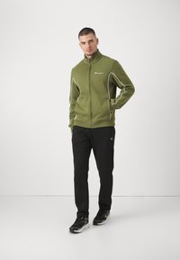 Zelená bunda na zips s bielym lemovaním, kombinovaná s čiernymi nohavicami s logom. Topánky sú čierne s neónovými akcentmi. Jednoduchý, športový dizajn.