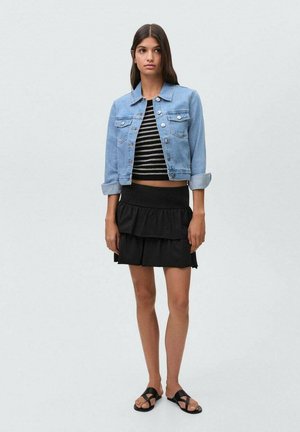 Veste en denim bleu clair au design court, crop top rayé noir et blanc, jupe à volants noire superposée et sandales noires.