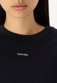 Felpa nera con colletto a coste e logo "Calvin Klein" ricamato in bianco sul davanti. Materiale morbido con una texture liscia.