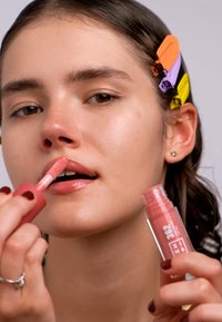 Gloss à lèvres dans un tube rose avec applicateur en forme de pied de doe, appliqué sur les lèvres. Le mannequin a les cheveux noirs avec des pinces colorées, mettant en valeur une peau lisse.