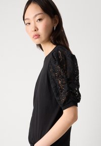 Μαύρη μπλούζα με φουσκωτά μανίκια από περίπλοκο μαύρο δαντέλα, σώμα από λείο ύφασμα, απλός λαιμός και εφαρμοστή γραμμή.