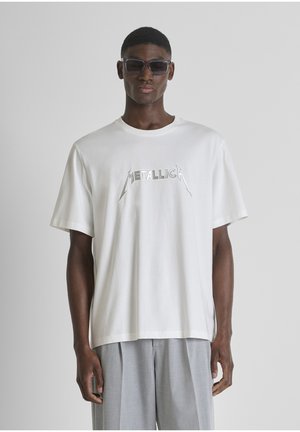 Uomo con occhiali da sole scuri, t-shirt bianca dei Metallica e pantaloni grigi chiari a pieghe su sfondo grigio uniforme.