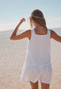 Biele šaty bez rukávov so štvorcovým výstrihom a rozšíreným lemom. Ľahká tkanina s mäkkou textúrou, nosené vonku na pláži.