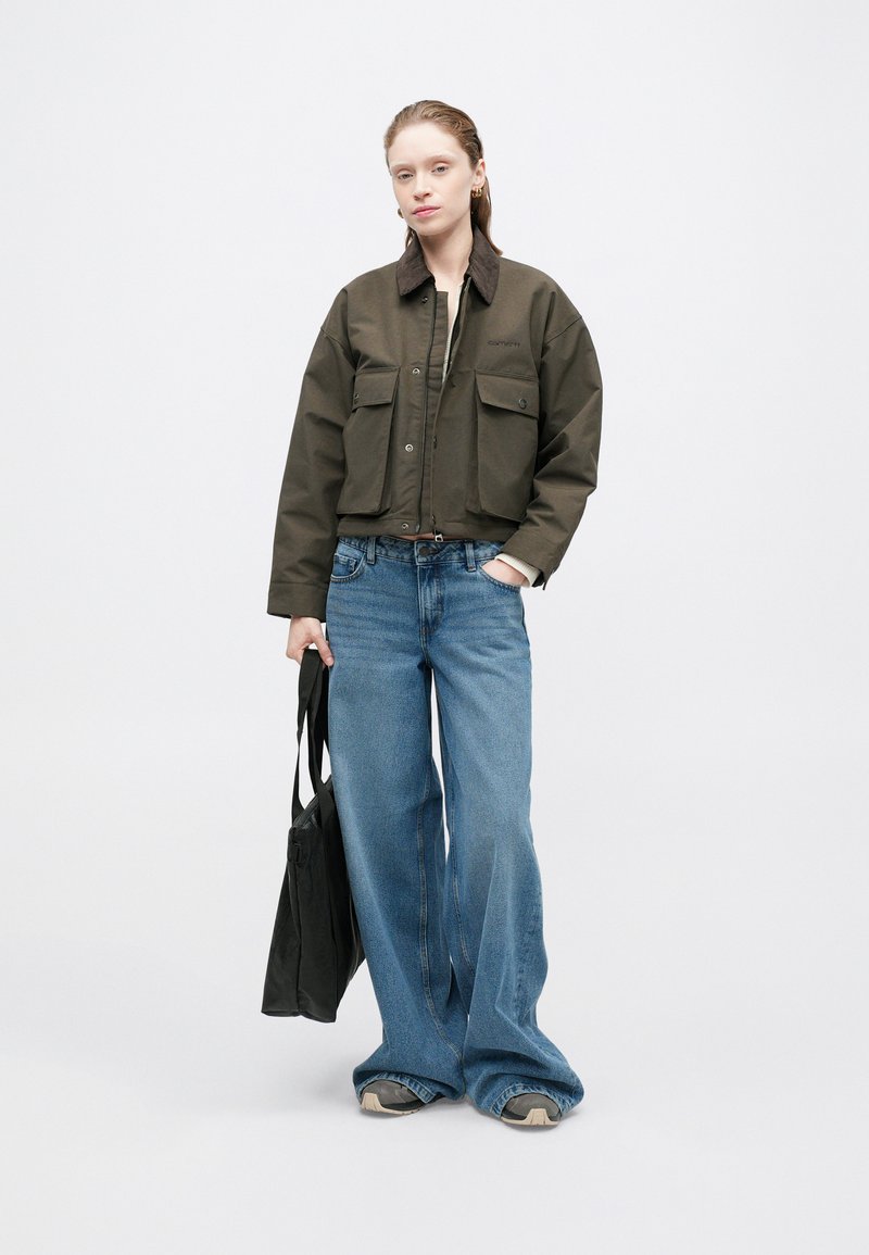 Gekürzte olivgrüne Jacke mit braunem Kragen, zwei Fronttaschen. Weite blaue Jeanshose, schwarze Tasche und graue Sneakers.