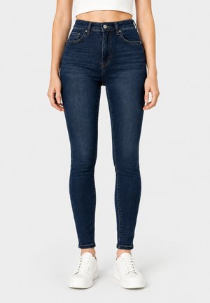 Kvinde iført mørkeblå skinny jeans og hvide sneakers, stående mod en ensfarvet lys baggrund.