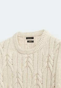 Beżowy sweter z warkoczy typu cable-knit z okrągłym dekoltem. Posiada teksturowany wzór z wyraźnymi warkoczami oraz ściągaczami na brzegach. Etykieta widoczna na dekolcie.