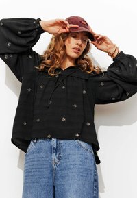 Blouse noire texturée avec des manches longues bouffantes et une petite broderie florale, assortie à un jean en denim bleu clair taille haute et une casquette bordeaux.