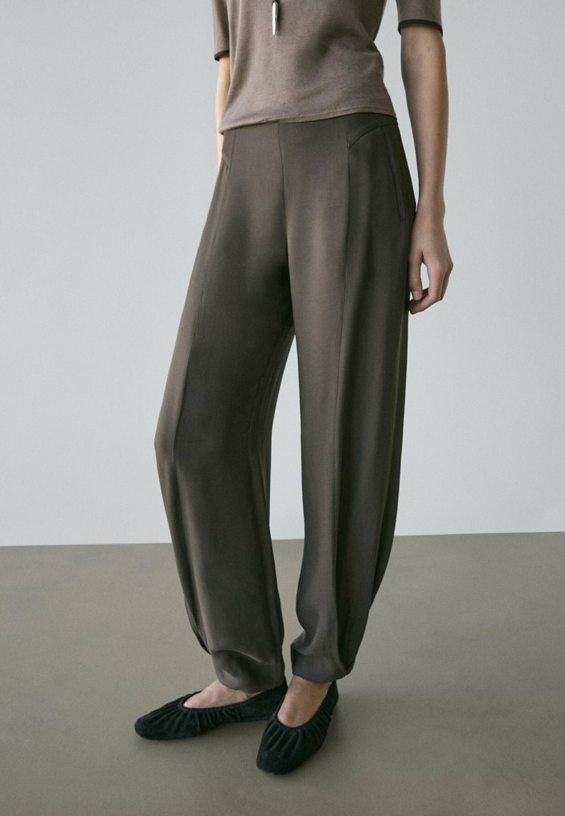 Frau trägt locker sitzende, dunkelbraune, hoch taillierte Hose mit Seitentaschen und schwarzen flachen Schuhen, steht auf einfachem Boden.