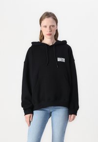 Hoodie noir en coton, design oversized avec une poche kangourou, capuche avec cordon de serrage, et un petit logo blanc sur la poitrine.