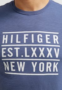Camiseta azul con texto blanco que dice "HILFIGER EST. LXXXV NUEVA YORK" en letras mayúsculas en el pecho.