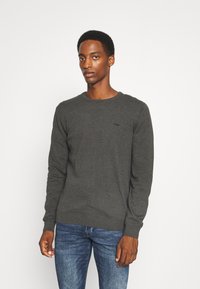 Grauer Pullover mit langen Ärmeln, rundem Ausschnitt, Strickstruktur und subtilen Logo-Detail auf der Brust. Kombiniert mit blauen Jeans.