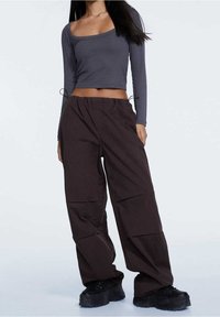 Vrouw in een grijze lange mouwen cropped top, losse zwarte broek met trekkoord, en zwarte chunky platform schoenen, staand tegen een effen achtergrond.