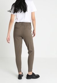Donkerbruine tapered broek met een elastische tailleband en achterzakken, gedragen met een wit T-shirt en zwarte schoenen, gezien van achteren.