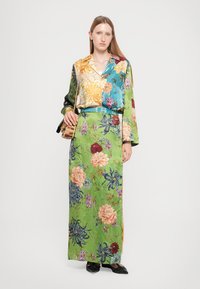 Camicia in seta floreale con tonalità blu, verde e oro abbinata a una lunga gonna verde con motivi floreali rosa e blu. Inclusi scarpe nere.