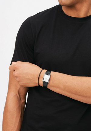Montre à bracelet en cuir noir avec un cadran rectangulaire argenté, présentée sur un poignet nu, associée à un bracelet noir fin. Fond de t-shirt noir.