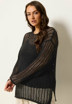 Frau mit langem braunem Haar, die einen schwarzen Strickpullover mit offenem Muster über einem hellen Oberteil trägt, vor einem schlichten hellen Hintergrund stehend.