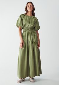 Abito maxi verde a maniche lunghe realizzato in tessuto leggero. Vita elastica e maniche a sbuffo. Le scarpe bianche perforate completano il look.
