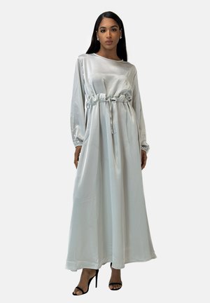 Vestido maxi de satén azul claro con mangas largas, cintura ceñida y detalle con cordón. Llevado con tacones de aguja negros. Textura suave.