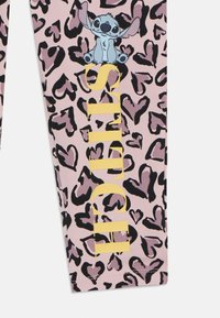 Rózsaszín leggings fekete és lila leopárdmintával, kék karakterrel és felszínes sárga felirattal, amelyen az áll "STITCH." Puha, rugalmas anyag.