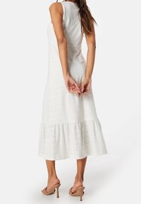 Bílé šaty s perforovanými detaily, bez rukávů, s délkou po kolena a volánkovým spodním lemem. Spárováno s hnědými lodičkami na vysokém podpatku. Pohled zezadu.