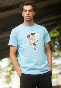 T-shirt en coton bleu clair mettant en avant un personnage de dessin animé dans une pose de football, portant un maillot rayé et une casquette, positionné sur un mannequin avec un arrière-plan extérieur.