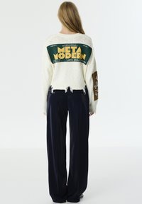 Pull en maille blanche texturé avec un motif graphique vert au dos portant la mention "META MODERN", associé à un pantalon large noir. Détails d'ourlet effiloché.