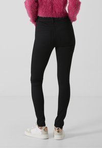 Person iført svarte tettsittende bukser, hvite joggesko med metalliske hæler, og en lys rosa pelskledd genser som står på en ensfarget lys bakgrunn.