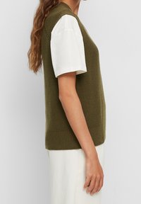 Oliwkowa dzianinowa kamizelka z teksturowanym wzorem, z okrągłym dekoltem i prążkowanym wykończeniem, noszona na białej koszuli z krótkim rękawem.