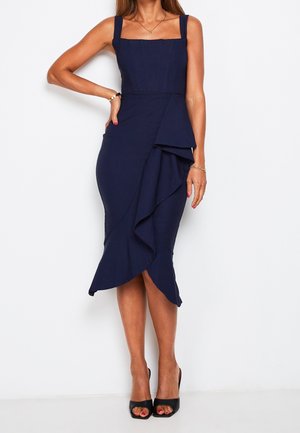 Vrouw draagt een marineblauwe mouwloze midijurk met vierkante halslijn en asymmetrisch ruchesdetail, gecombineerd met zwarte open teen hakken.