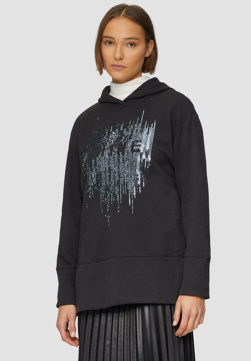 Czarna bluza z kapturem pokryta wzorem z cekinów w pionowym układzie. Posiada luźny krój oraz ściągacze na mankietach, zestawiona z plisowaną spódnicą.