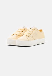 Gelbe Leinensneaker mit weißen Gummisohlen, runder Zehenpartie, fünf Ösen und weißen Akzenten auf der Zunge, aus einem strukturierten Stoff gefertigt.