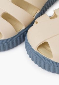 Lightweight sandals made of beige plastic with a blue ribbed sole. Features open toe and side vents for breathability.