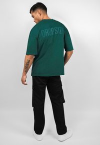 Grünes Baumwoll-T-Shirt mit Rundhalsausschnitt und kurzen Ärmeln, auf der Rückseite mit einem großen, strukturierten "DROPSIZE"-Druck; kombiniert mit schwarzen Cargo-Hosen.