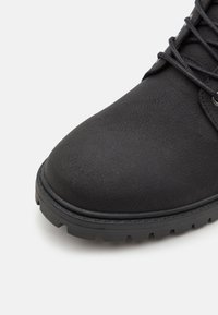 Botín negro de tobillo hecho de material texturizado, con puntera redondeada, suela resistente con agarre y cordones gruesos en la parte superior.