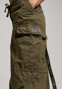 Olijfgroene cargobroek met een taillekoord, meerdere zakken en een label met de tekst "PARA PANT 2003." Gevoerd, lichtgewicht stof.